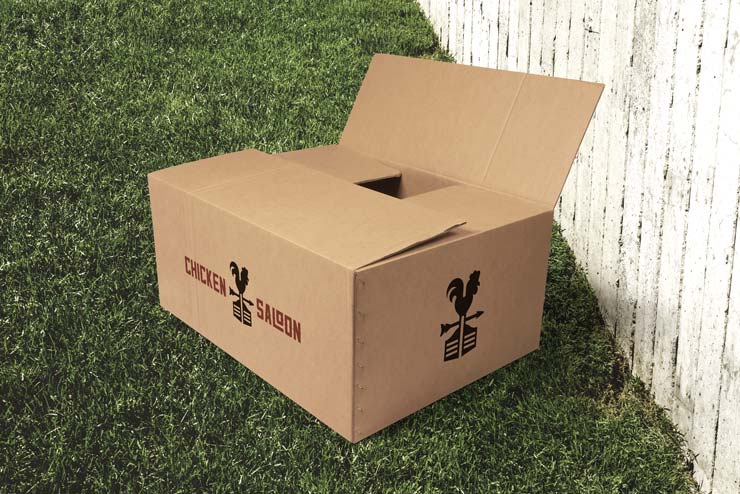 A corrugated cardboard box with Chicken Saloon logos on it, partially opened on a grassy lawn